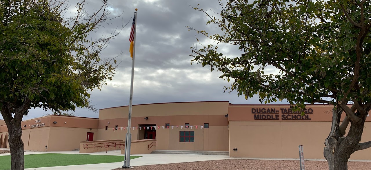 A low-angle, eye-level, panoramic photo capturing the front facade of a single-story school building. The building is painted in various shades of tan and brown, characteristic of Southwestern architecture, under an overcast sky.  A prominent flagpole stands in the foreground, displaying the American flag with the New Mexico Zia symbol below it, indicating the location is New Mexico.  The school's name, "DUGAN-TARANGO MIDDLE SCHOOL," is visible in dark lettering on the right side of the central, curved section of the building. To the left, another sign with smaller text reads "CENTRAL ELEMENTARY SCHOOL," suggesting a dual-purpose or co-located facility.  In front of the school, there's a well-maintained green lawn area, a paved walkway leading to the entrance, and a concrete ramp for accessibility. Two trees with sparse foliage frame the building on either side, adding to the outdoor setting. The immediate foreground has some gravel landscaping on the right.