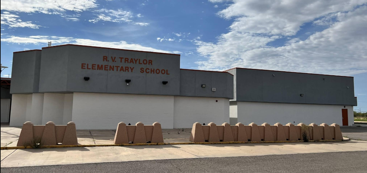 A long, single-story school building under a partly cloudy sky. The building has a modern, somewhat utilitarian design with sections painted in light gray and white. Above the main entrance, the name "R.V. TRAYLOR ELEMENTARY SCHOOL" is prominently displayed in orange-red letters on a gray band. In the foreground, a row of light brown, concrete barrier blocks with rounded tops extends across the width of the image, separating a paved area in front of the school from what appears to be a road or parking lot in the immediate foreground. Some small weeds are visible around the base of the concrete blocks. The surrounding area appears flat and somewhat arid, typical of the landscape in Deming, New Mexico.