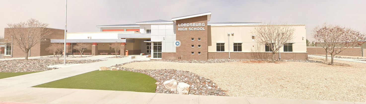 A wide, eye-level panoramic photo of Lordsburg High School, a modern, multi-building campus with a blend of brick and light-colored stucco facades under a bright sky.  The central building features prominent brickwork and a distinct architectural design with varying rooflines. The school's name, "LORDSBURG HIGH SCHOOL," is clearly displayed in large, white letters on a light-colored section above the main entrance. A school emblem or logo is visible on the brick wall near the entrance.  To the left of the main building, there's an open-air covered walkway leading to another section of the school, with picnic tables visible underneath.  The landscaping in the foreground and around the buildings consists of low-water desert plants, gravel, and some decorative rocks, with patches of artificial turf near the walkways. Several deciduous trees, currently without leaves, are scattered throughout the grounds. The overall impression is a well-maintained and contemporary educational facility in an arid environment.