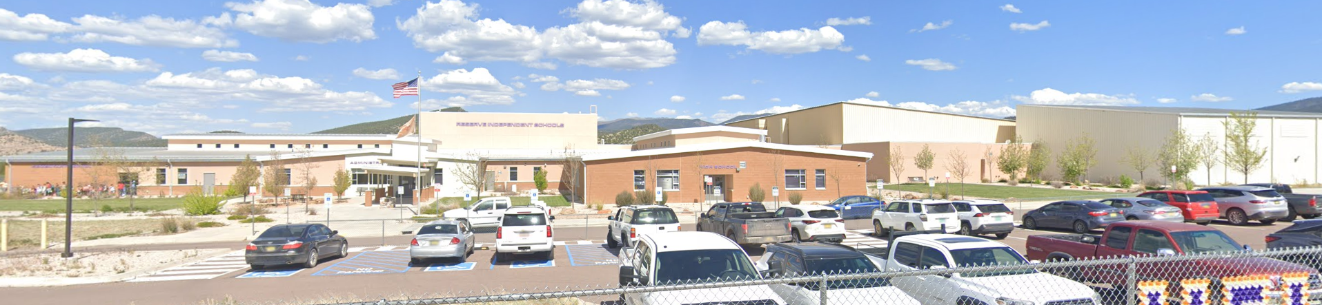 A wide, panoramic view of a large school campus, likely in a mountainous or hilly region, under a bright blue sky with scattered white clouds. The school buildings are primarily light-colored (tan or beige) with some brick accents and flat roofs.</p>
<p>In the center-left, an American flag flies from a flagpole. The name "ESPANOLA VALLEY PUBLIC SCHOOLS" is visible on the main building's facade. The campus appears to consist of multiple connected buildings of varying heights and designs, suggesting different wings or sections of a large educational complex.</p>
<p>In the foreground, there is a paved parking lot filled with various cars, indicating an active school environment. A chain-link fence separates the viewer from the parking lot. In the background, rolling hills or mountains are visible, covered with trees, which contributes to the scenic setting of the school.