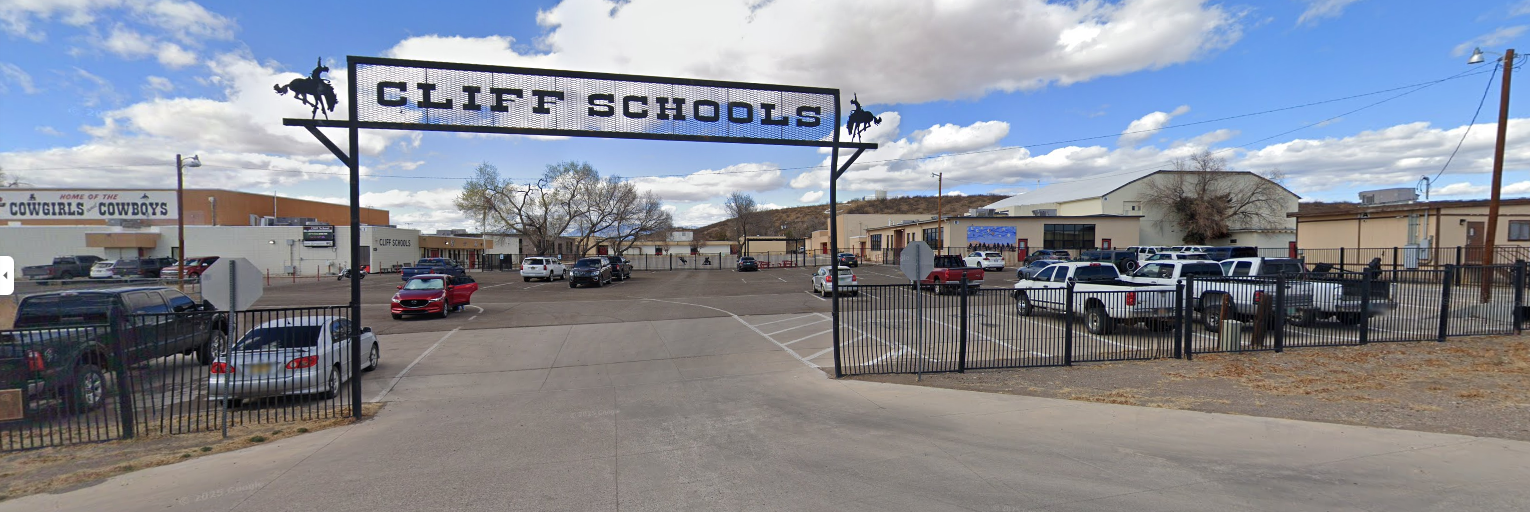 A wide, panoramic view of the entrance to "CLIFF SCHOOLS." A large, rustic-style metal archway spans across the main entrance, with the words "CLIFF SCHOOLS" prominently displayed at the top in bold, white letters. Silhouettes of two horses, one on each side, adorn the archway.  Beyond the archway, a paved drive leads into a large school campus with several buildings visible. The buildings appear to be a mix of single and two-story structures, mostly in light colors like white or tan. A large building with a sloped roof and a basketball hoop is visible on the right side of the campus. Another building on the far left has a sign that reads "HOME OF THE COWGIRLS - COWBOYS," indicating the school's mascot.  The campus includes multiple parking areas with various cars and pickup trucks. The ground is a mix of asphalt and arid-looking dirt or gravel, consistent with a dry, possibly desert, environment. The sky is partly cloudy with patches of blue. In the distance, hills or mesas can be seen. The overall impression is a large, spread-out school complex in a rural or semi-rural setting.