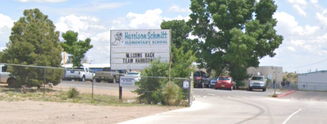 A panoramic outdoor shot of the entrance to Harrison Schmitt Elementary School on a bright, partly cloudy day.  In the center foreground, a large white wooden sign with blue text reads "Harrison Schmitt ELEMENTARY SCHOOL" at the top, along with a cartoon mascot that appears to be a badger or similar animal. Below this, in black handwritten-style letters, it says "WELCOME BACK TEAM HARRISON." The sign is surrounded by some green bushes and dry, arid ground.  A chain-link fence runs across the left side of the image, behind which some parked white pickup trucks are visible. The ground is dry and dusty.  To the right of the sign, a paved driveway curves into the school grounds. Several vehicles, including a red car and a silver SUV, are visible on the paved area. In the background, there are various school buildings, mostly single-story and light-colored, with some dark green trees providing shade. The sky is blue with fluffy white clouds. The overall impression is a welcoming, established elementary school in a dry, possibly Southwestern, environment.