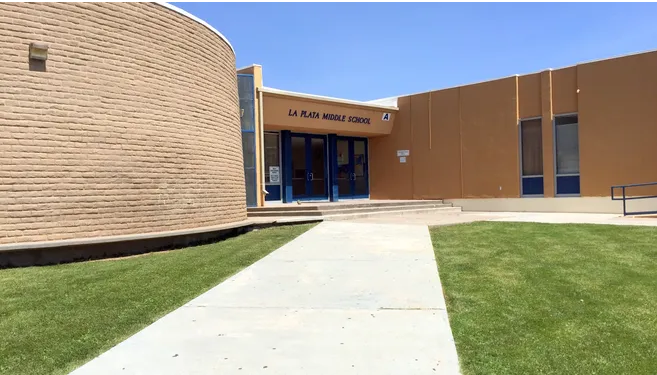 A head-on, slightly low-angle shot of the main entrance to La Plata Middle School on a bright, sunny day. The school building is primarily a muted orange color with a large, curving section of light brown brick on the left. A wide concrete walkway leads from the foreground towards the double blue doors of the entrance. Above the doors, the words "LA PLATA MIDDLE SCHOOL" are visible. Green grass lawns flank the walkway on both sides, and the sky above is a clear, light blue.