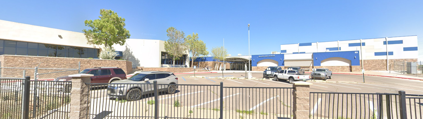 A panoramic view of Silver High School under a clar blue sky. The school building features a mix of architectural styles, with sections of light-colored stucco and blue metal siding. On the left, there's a curved beige building with large, dark-tinted windows, and a stone wall runs along the bottom. In the center, a covered walkway leads to the main entrance, and a sign above reads "DEMING HIGH SCHOOL." Several cars are parked in the lot in front of the school. A black metal fence with a dieamond pattern is in the foreground, separating the viewer from the parking lot and school.