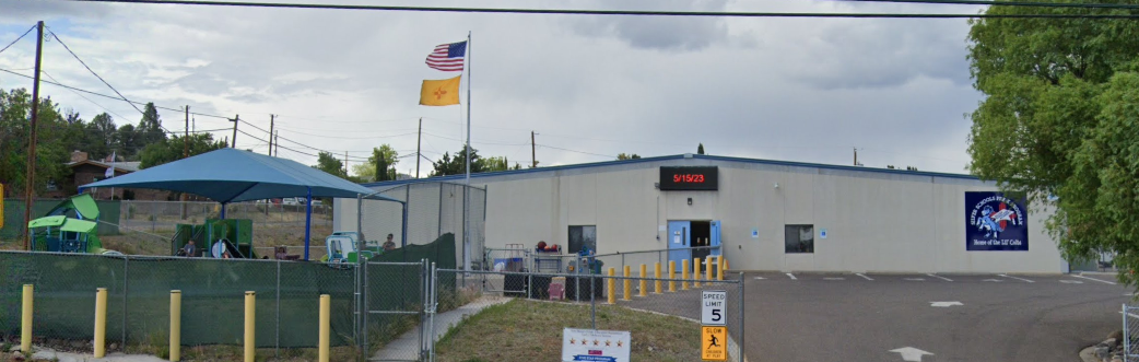 A wide, outdoor shot of the Silver Scholars academy on a cloudy day. The main building is a light-colored, single-story structure with blue trim around the windows and doors. An electronic sign above the entrance displays "9/15/23." To the left of the building, there's a playground area with a large blue shade canopy and green play equipment. Two flagpoles stand in front of the building, flying the American flag and the New Mexico state flag. A paved parking lot with a "SPEED LIMIT 5" sign and yellow bollards is in the foreground. On the right side of the building, there's a logo that reads "Silver Scholars Academy Home of Lil Colts."