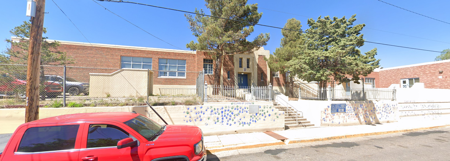 A slightly elevated, wide-angle shot of a school building on a sunny day. The main building is made of light brown brick, with a central section that appears to be stucco or concrete in a lighter color, featuring a set of blue double doors at the entrance. Steps with white railings lead up to the entrance. In front of the school, there's a light-colored wall with a mosaic pattern of blue and white tiles. A chain-link fence runs along the property line, behind which are some trees and bushes. A large red pickup truck is parked on the street in the foreground, partially obscuring the lower left of the image. A sign is visible on the right side of the mosaic wall.