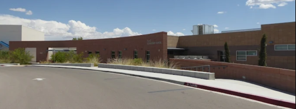 A low-angle, panoramic shot of T or C Elementary School on a sunny day with a blue sky and scattered white clouds. The school building has a modern design with a mix of reddish-brown and lighter brown facades, and a distinctive curved section in the center. The words "Tor C ELEMENTARY SCHOOL" are visible on this curved part. There are several windows and some landscaping with arid-climate plants along the front of the building. A paved driveway or parking area is in the foreground, with a white arrow indicating direction and a red curb with "NO PARKING" written on it.