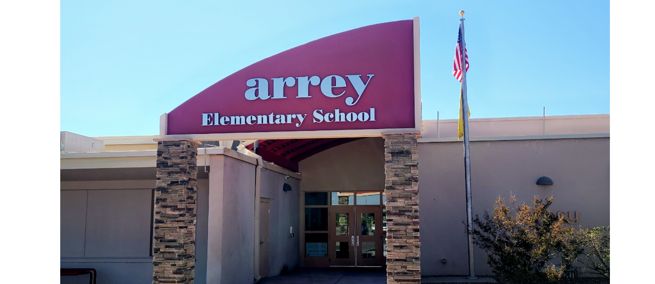 A sunny, head-on shot of the entrance to Arrey Elementary School. The main sign above the entrance is a large, curved burgundy canopy with the word "arrey" in large, stylized white letters and "Elementary School" in smaller white letters beneath it. The entrance itself is set back, with double doors visible, and is flanked by columns with stacked stone facing. To the right of the entrance, an American flag flies from a flagpole. The sky is a clear, bright blue.