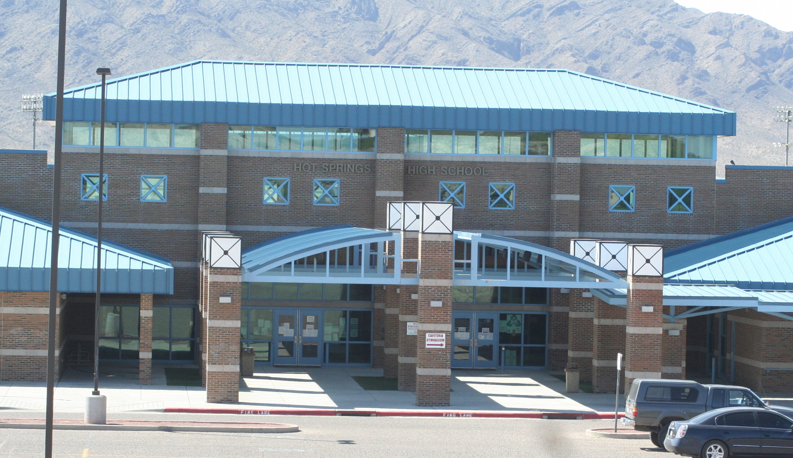 A direct, head-on shot of the entrance to Hot Springs High School, with mountains visible in the hazy background. The school building features a prominent light blue metal roof and light brown brick walls. The main entrance has a large, arched blue canopy supported by brick columns. Above the entrance, "HOT SPRINGS HIGH SCHOOL" is written on the building. Several windows are visible, some with blue accents forming an 'X' pattern. A "VISITOR PARKING" sign with an arrow is visible near the entrance. In the foreground, there's a paved area with "FIRE LANE" painted on the curb, and a dark blue SUV is partially visible on the right.