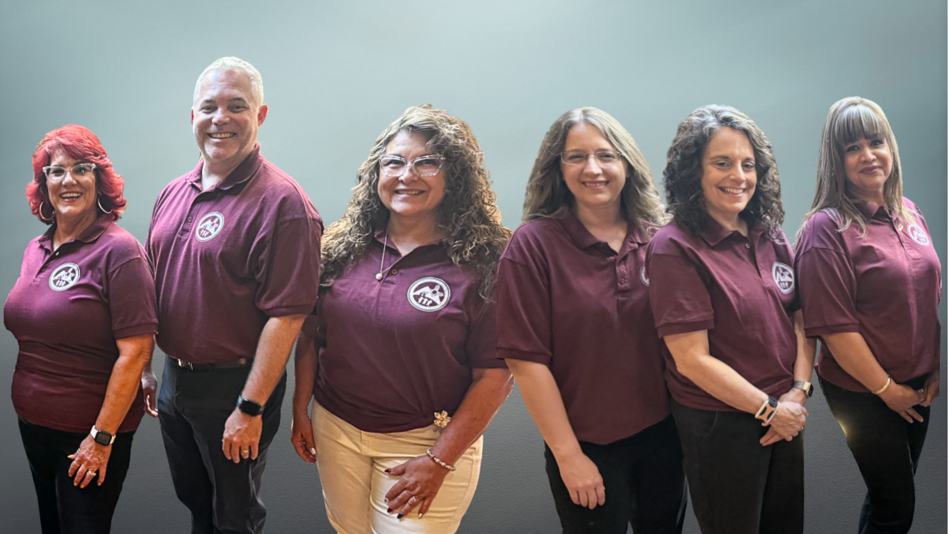 <br />
A group of six smiling people stand in a line against a gray background. They are all wearing matching maroon polo shirts with a circular logo on the left chest. From left to right, the people are: a woman with short reddish-brown hair and glasses, a man with short gray hair and a clean-shaven face, a woman with long curly brown hair and glasses, a woman with long straight brown hair and glasses, a woman with long curly brown hair, and a woman with straight brown hair and bangs.