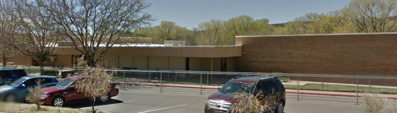 A wide, panoramic, eye-level shot of Jose Barrios Elementary School on a sunny day. The school building is primarily a light tan color, with a long section of brick wall on the right side. The architecture appears functional, with flat roofs.  In the foreground, there's a paved parking lot with a few cars visible, including a dark blue sedan and a maroon SUV. A red curb lines the edge of the parking lot, beyond which a chain-link fence runs across the width of the image, separating the parking area from the school grounds.  Behind the fence, there's a grassy area and several bare deciduous trees, suggesting the photo was taken in late winter or early spring. In the background, beyond the school, a dense line of trees with green foliage indicates a forested or heavily vegetated area. The sky is clear and blue.