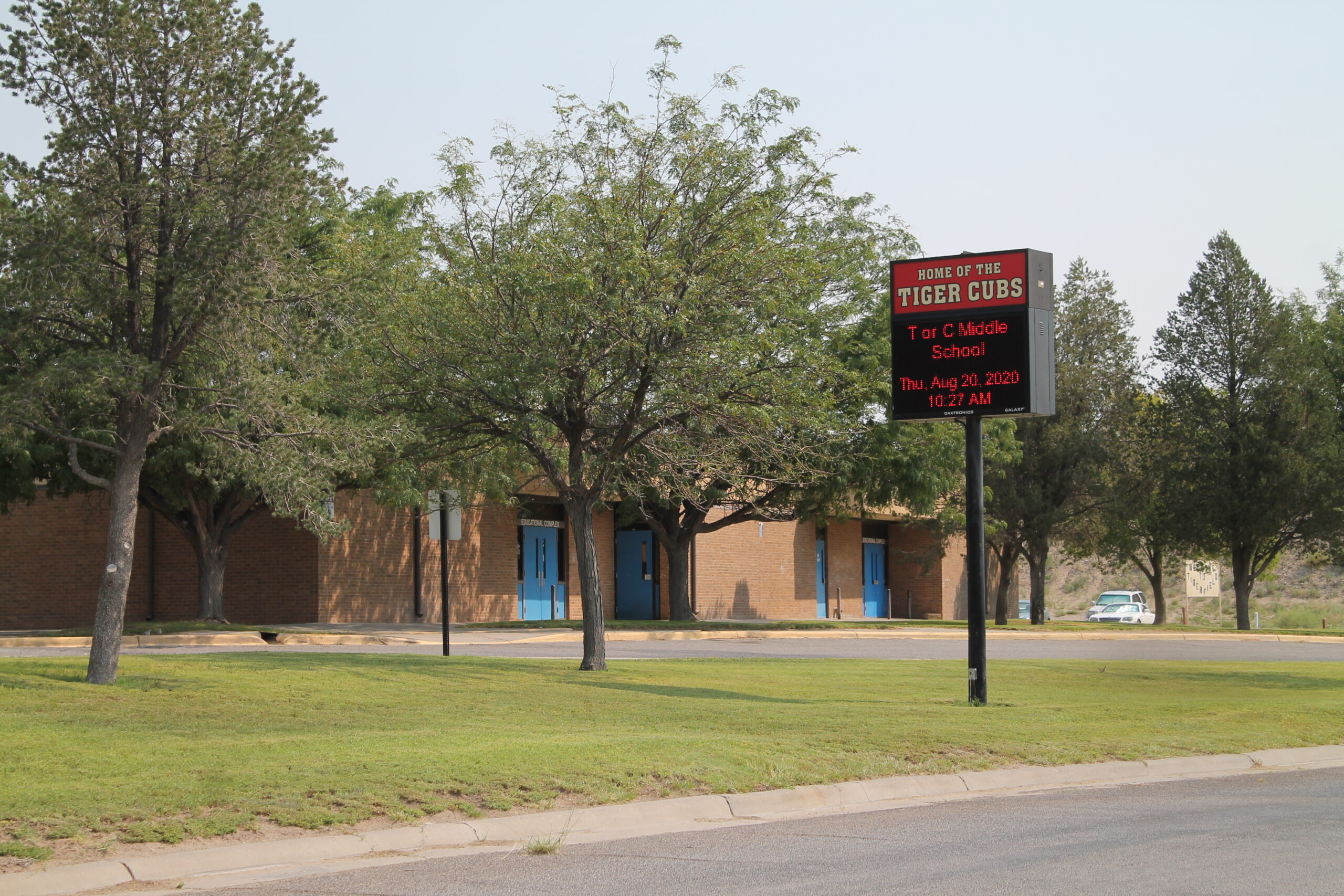 A sunny, wide exterior shot of Tor C Middle School with a prominent digital sign. The school building, made of light brown brick, is visible behind a grassy lawn and several mature trees. The building has multiple light blue double doors. On the right side of the image, a large black digital sign displays "HOME OF THE TIGER CUBS," "Tor C Middle School," and "Thu. Aug 20, 2020 10:27 AM." A road curves in the foreground, and a white car is partially visible in the distance to the right of the sign. The sky is hazy.