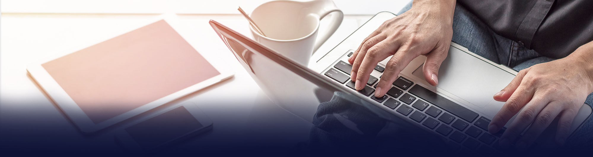 person sitting with laptop on lap with phone, tablet and mug next to them