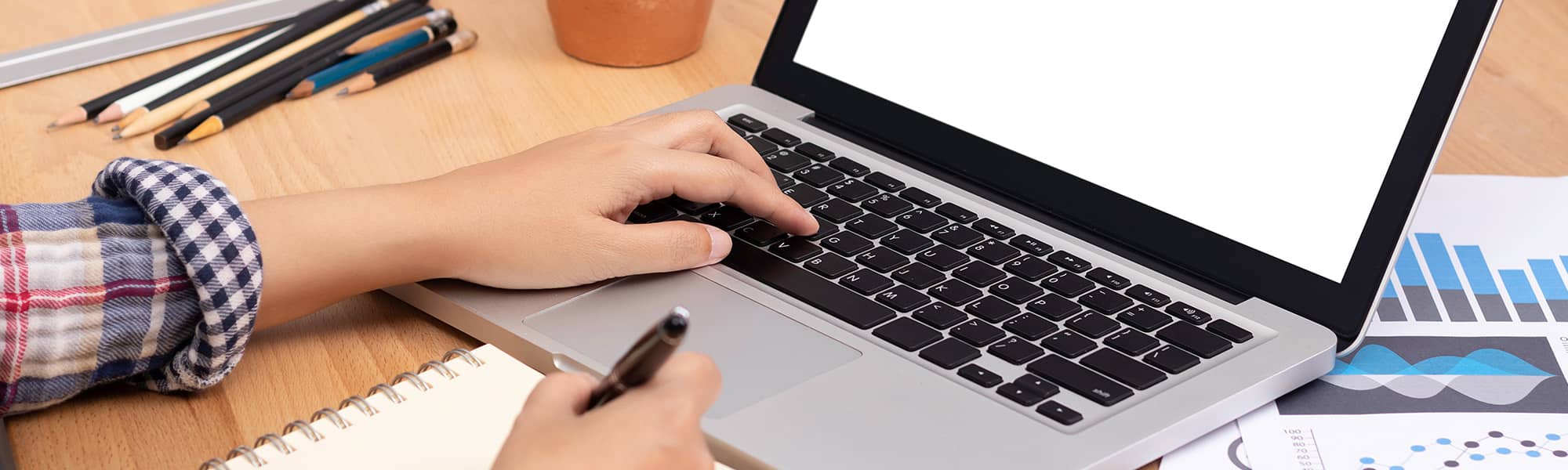 student typing on a laptop while writing in a notebook