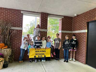 students standing in front of a window holding a banner that reads Walking School Bus