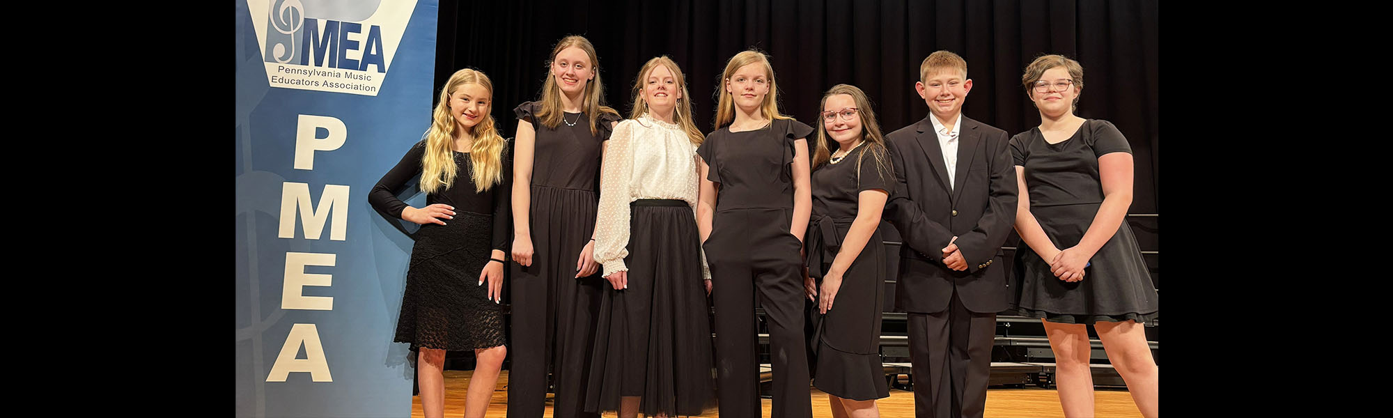 Group of happy students dressed in formal attire next to a Pennsylvania Music Educators Association (PMEA) banner