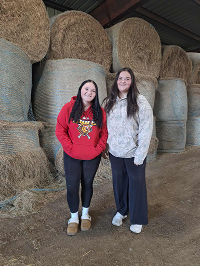 Two girls posing inside a facility filled with large rolls of hay