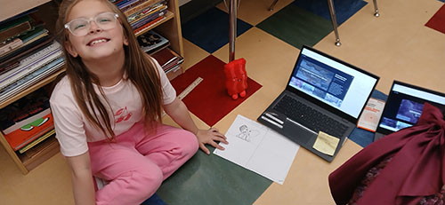 Happy third grade student sitting on the classroom floor with a laptop