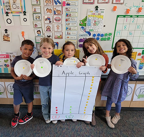 Group of happy kindergarten students holding up their apple graphing project in the classroom