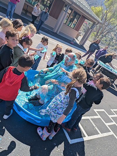 Principal McNelis sits on a chair in a kiddie pool while children pour blue slime on him.