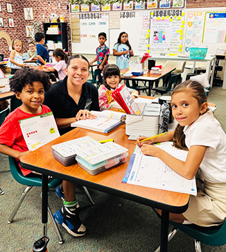 Happy adult helping elementary students at table in the classroom